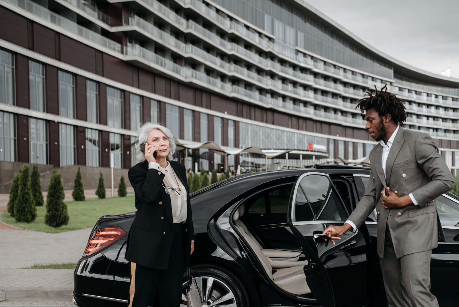 A private driver in a suit holds the rear door of a black car open for a passenger outside a modern hotel, the kind of service you get when you hire a private driver