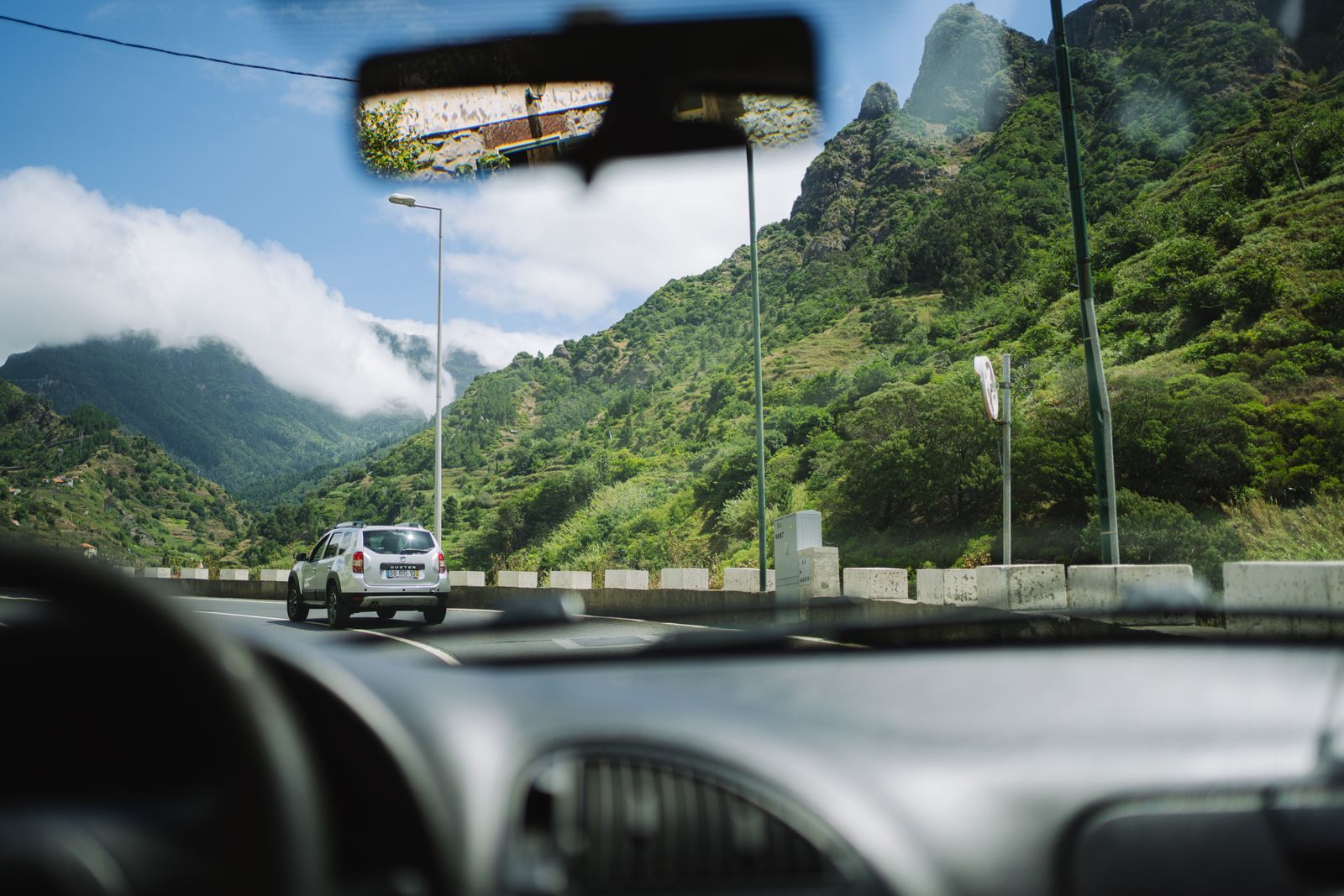 View through a car windshield of a mountain road with green slopes and a car ahead