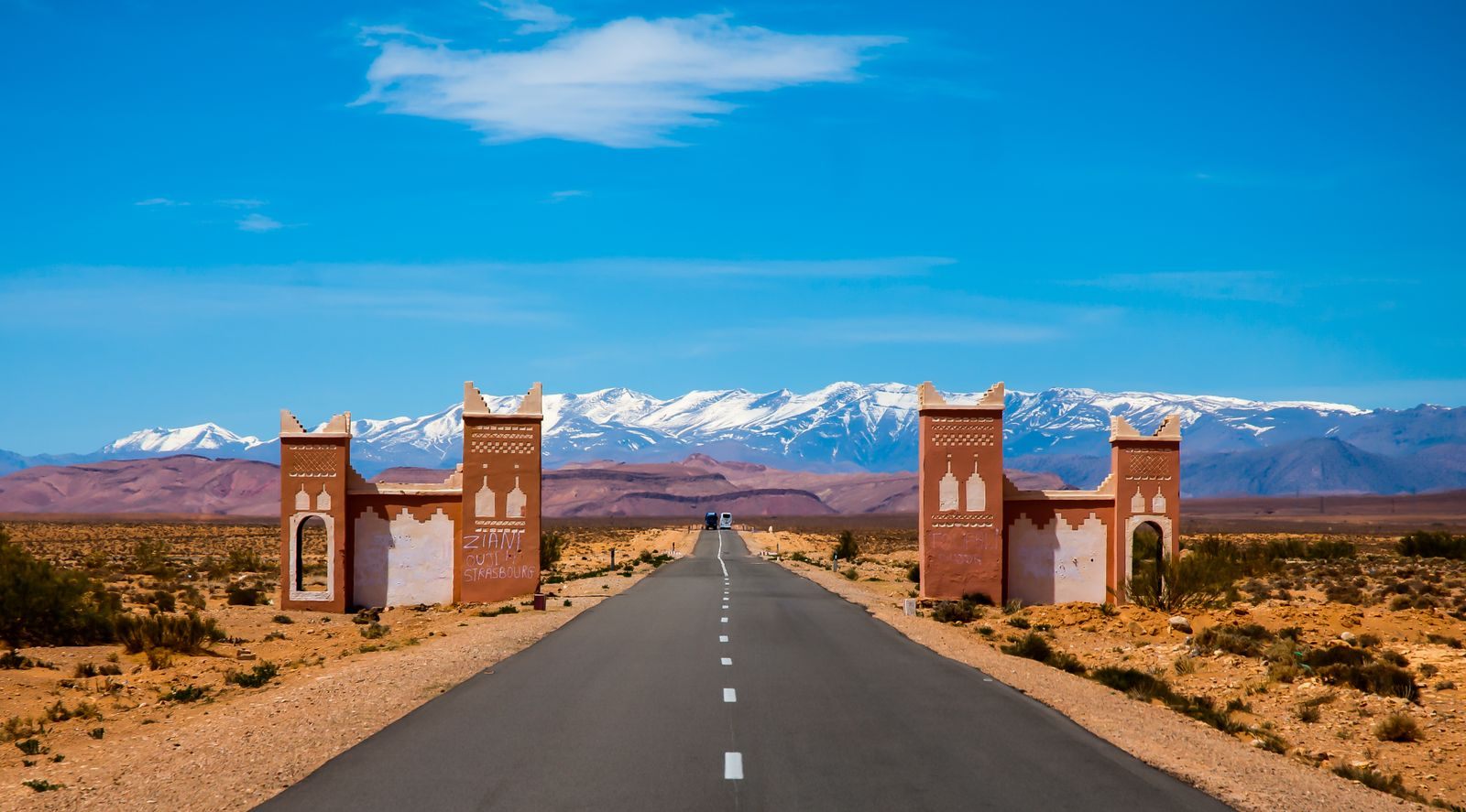 Straight desert road passing between two earthen gate towers toward snow-capped mountains